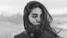 Woman with windblown hair looking introspective in black-and-white. Soft blurred background and somber mood. Unsplash watermark bottom left.