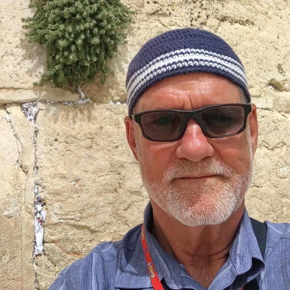 Man with sunglasses and striped cap smiles in front of ancient stone wall. Red lanyard with "GATE 1" text. Sunny day. Scrub plant visible.