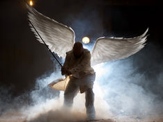 A person with large white wings and a sword kneels in a misty, dimly lit space, conveying a dramatic and mysterious mood.