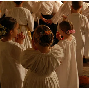 Children in white dresses with floral hairpieces clap in a dimly lit room, creating a warm, joyful atmosphere.