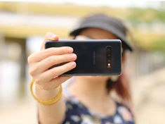 Person taking a selfie with a smartphone, wearing a floral top and cap. Blurry background, focus on phone. Mood is casual.