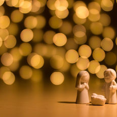Three white figurines depict a nativity scene on a smooth surface with a warm bokeh background of golden lights, creating a peaceful mood.