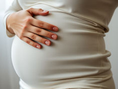 Close-up of a pregnant woman in a cream dress gently resting her hand on her belly by a bright window.