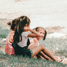 Two girls sitting on grass, with the younger girl smiling joyfully. Bright, sunny setting with blurred background. Calm and cheerful mood.