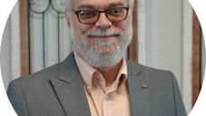 Smiling man with glasses and a beard in a gray suit and peach shirt, standing indoors with decorative framed background.