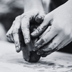 Hands shaping clay on a table. Fingers are coated with mud, suggesting a focus on creativity. The scene is in grayscale, adding a calm mood.