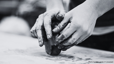 Hands shaping clay on a table. Fingers are coated with mud, suggesting a focus on creativity. The scene is in grayscale, adding a calm mood.