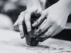 Hands shaping clay on a table. Fingers are coated with mud, suggesting a focus on creativity. The scene is in grayscale, adding a calm mood.