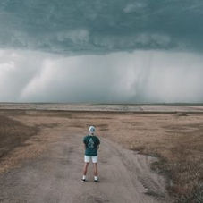 A person in shorts stands on a dirt road facing a stormy sky, watching a lightning bolt. Vast field under ominous dark clouds.