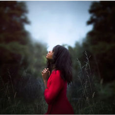Woman in a red dress stands in a field, gazing upwards with hands clasped, surrounded by tall grass and trees under a cloudy sky.