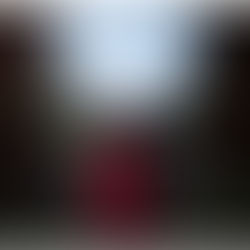 Woman in a red dress stands in a field, gazing upwards with hands clasped, surrounded by tall grass and trees under a cloudy sky.