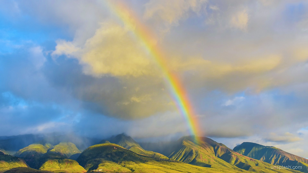 Rainbow arcs over lush green mountains under a cloudy sky. Soft sunlight illuminates the scene, creating a serene, uplifting mood.