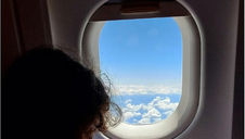 Child gazes out airplane window at blue sky and clouds. Interior wall is gray. Scene evokes calm and curiosity.