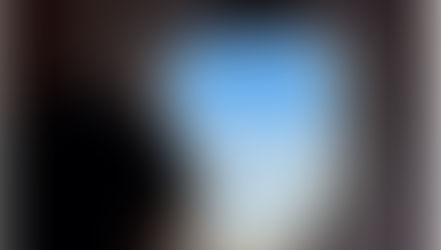 Child gazes out airplane window at blue sky and clouds. Interior wall is gray. Scene evokes calm and curiosity.