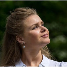 Woman with long hair looks up, smiling slightly. Wearing a white top and earrings. Blurred green background suggests an outdoor setting.