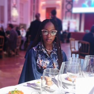 Woman with glasses sits at a formal dinner table in a softly lit pink room, with wine glasses, bread, and other guests blurred behind.