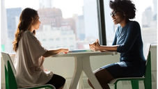 Two women sit across a table in a bright office with city views, engaged in conversation. One wears a blue dress, the other light casual attire.
