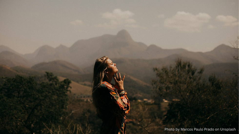 Woman in a colorful dress stands in a peaceful landscape, eyes closed and hands near her face, with mountains in the hazy background.