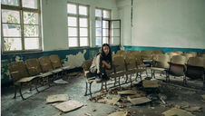 Woman crouches on a chair in a dusty, abandoned classroom with broken seats and peeling paint. Mood is desolate. Light streams in.