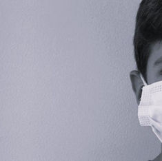 Young boy wearing a white face mask, partially visible against a plain light background. The image has a calm, muted tone.