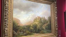 A landscape painting in a detailed gold frame hangs on a red wall. It depicts a lush valley with a castle on a hill under a cloudy sky.