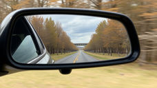 Car's side mirror reflects a tree-lined road under a cloudy sky, with fall foliage and a distant bridge, evoking a serene mood.