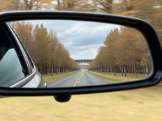 Car's side mirror reflects a tree-lined road under a cloudy sky, with fall foliage and a distant bridge, evoking a serene mood.