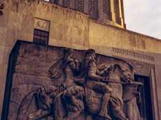 Stone relief of two horseback riders, "Lewis and Clark," on a building facade with "State of Oregon" text. Warm evening light, historic mood.