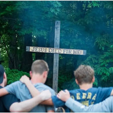 Four people with arms around each other face a wooden cross with "JESUS DIED FOR ME!" text. Background is lush green forest, creating a serene mood.