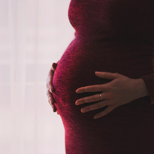 Pregnant woman in a burgundy dress holding her belly, standing against a sheer, light curtain. Light creates a peaceful, intimate mood.