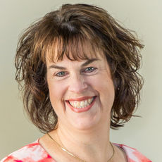 Smiling woman with short, curly brown hair and a red patterned top against a plain background, conveying warmth and friendliness.