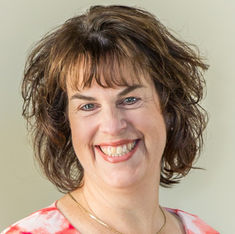 Smiling woman with short, curly brown hair and a red patterned top against a plain background, conveying warmth and friendliness.