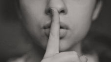Close-up of a person holding a finger to their lips, signaling silence. Grayscale tones and soft focus create a calm, quiet mood.