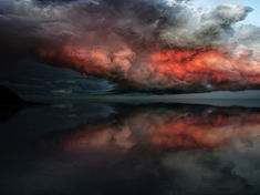 Dramatic clouds with red hues reflect on a calm, dark water surface. A cloudy sky casts a moody atmosphere, with a silhouette of land.