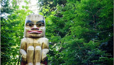 Carved wooden totem pole with a stern face among lush green forest foliage. Bright sunlight filters through the leaves, creating a serene mood.