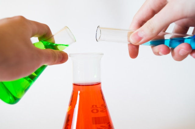 Hands pour green and blue liquids from test tubes into a red solution in a beaker, set against a plain white background.