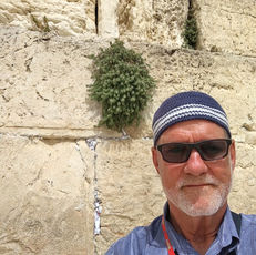 Man with sunglasses and striped cap smiles in front of ancient stone wall. Red lanyard with "GATE 1" text. Sunny day. Scrub plant visible.