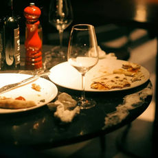 Empty wine glasses, plates with pizza remnants, red pepper grinder, and crumpled napkins on a dimly lit table create a cozy, used setting.