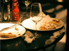 Empty wine glasses, plates with pizza remnants, red pepper grinder, and crumpled napkins on a dimly lit table create a cozy, used setting.
