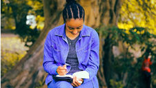 A person in a blue jacket sits on a bench under a tree, writing in a notebook. The background is a sunlit park with blurred foliage.