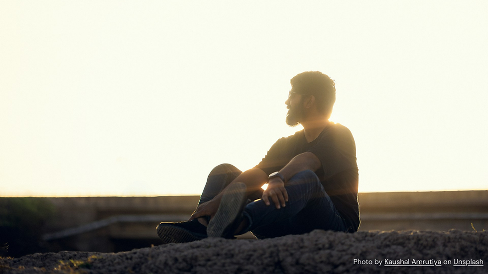 A man in a black shirt sits on a rock, gazing into the distance during sunset. The background is bright and serene.
