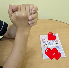 AppaPrayer clasping hands over a wooden table beside a Korean booklet reading 소망부 주보, in a calm, supportive scene.