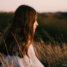 Young woman with long hair sitting in a grassy field, gazing into the distance at sunset. The mood is calm and reflective.