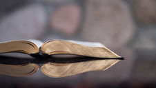 Open Bible lying on a glossy surface reflects faintly. Soft-focus stone wall in the background. Calm and serene atmosphere.