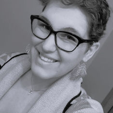 Woman smiling in black glasses and patterned cardigan, wearing a necklace and large earrings. Black and white image with a cheerful mood.