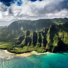 Aerial view of a lush, green mountain coastline meeting turquoise ocean waters under a partly cloudy sky, creating a serene and dramatic landscape.