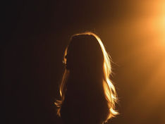 Silhouetted woman with long hair backlit by warm orange light against a dark background, creating a dramatic glow.