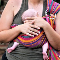Woman holding a baby in a colorful striped sling against a brick wall. The baby's head rests under the adult's hand, evoking calm and care.