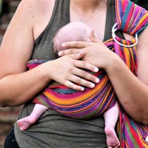 Woman holding a baby in a colorful striped sling against a brick wall. The baby's head rests under the adult's hand, evoking calm and care.