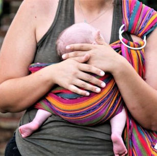 Woman holding a baby in a colorful striped sling against a brick wall. The baby's head rests under the adult's hand, evoking calm and care.